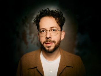 junger Mann mit kurzen, dunklen, lockigen Haaren, dunklem Bart und Goldrandbrille